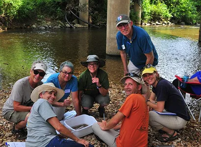 Seven adults kneel and sit on rocks by a stream under a bridge, smiling at the camera. They wear hats and casual outdoor clothing, and gather around white trays, suggesting a nature or science activity.