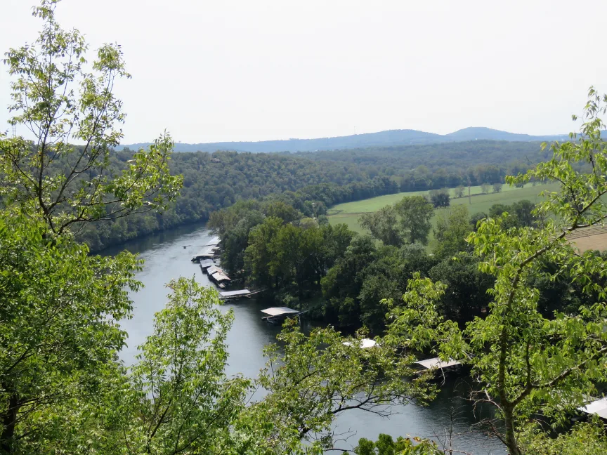 A river winds through a lush, green landscape with trees on both sides and several boat docks along the water. Rolling hills and open fields are visible in the background under a hazy sky.