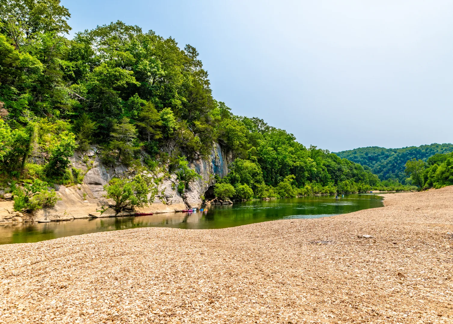 A calm river curves beside rocky cliffs and dense green trees, with a wide, pebbled shoreline in the foreground and forested hills in the background under a clear sky.