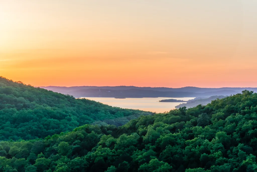 A scenic view of lush green hills with a lake in the distance, under a colorful sunrise or sunset sky with orange and pink hues.