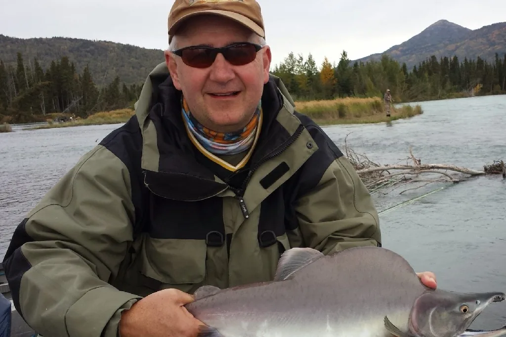 A smiling man in a jacket and cap holds a large fish by a river, with trees and mountains in the background.