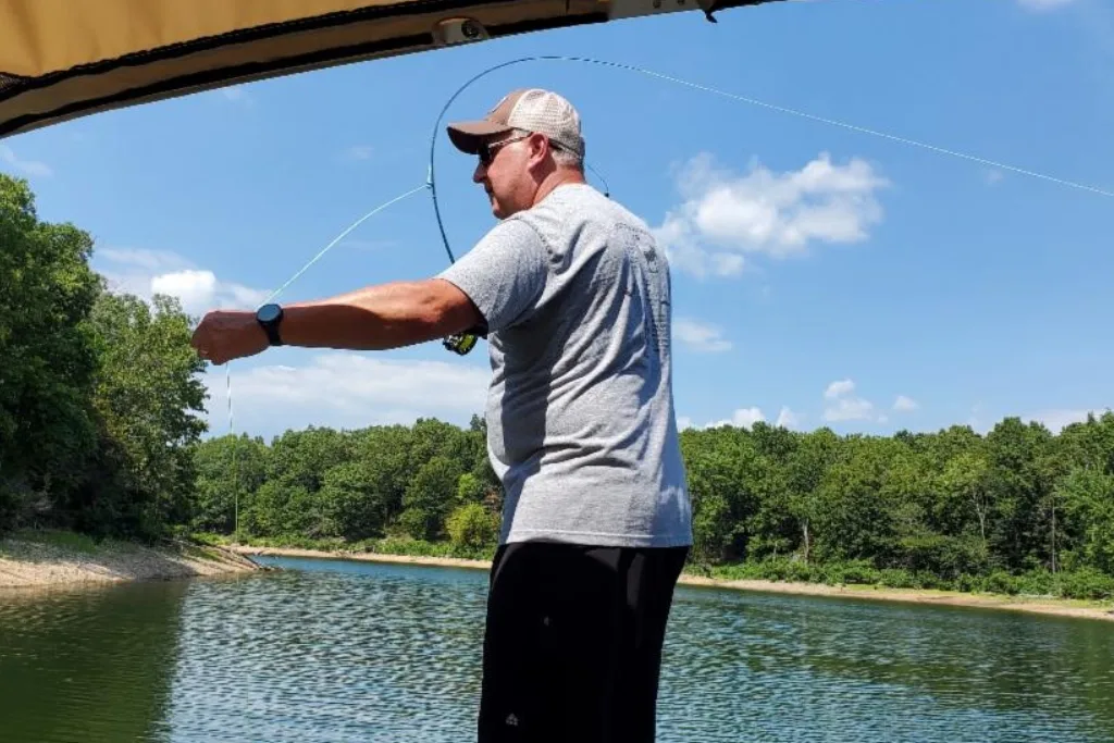 A man wearing a gray t-shirt, black shorts, and a baseball cap is fishing on a boat, holding a bent fishing rod over a calm lake surrounded by trees under a blue sky with scattered clouds.