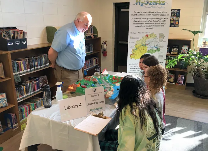 A man stands beside a table labeled Library 4 and Jerry Harmon, speaking with three seated girls. The table has educational materials, and bookcases and a H2Ozarks informational display are visible in the background.