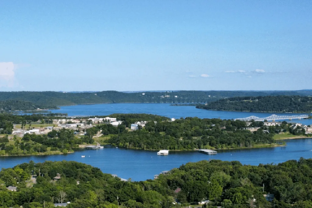 A scenic aerial view of a lake with small boats, tree-covered islands, a bridge, and buildings surrounded by lush green hills under a clear blue sky.