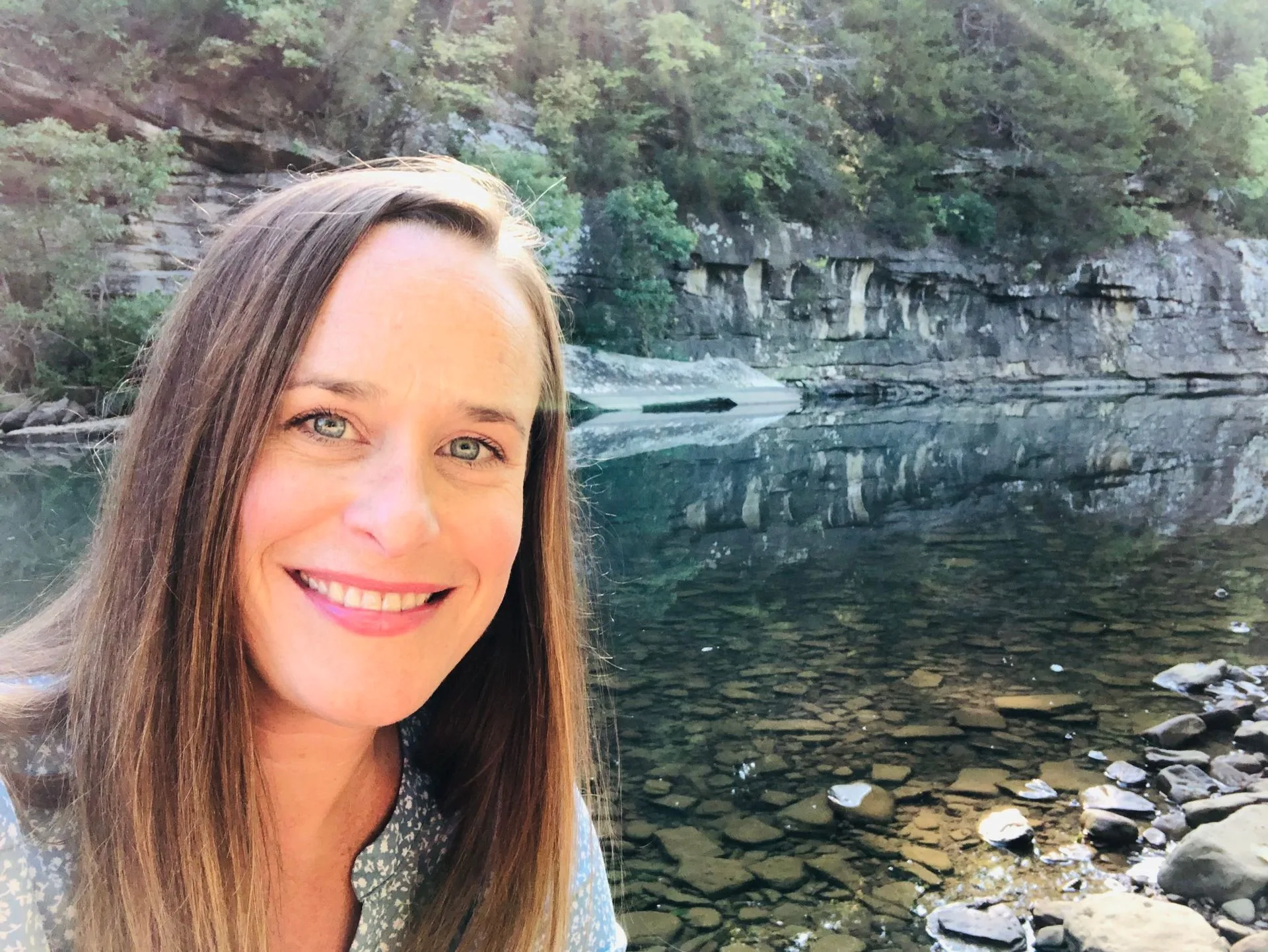 A woman with long brown hair smiles at the camera while standing near a rocky riverbank, with clear water and green trees reflecting in the background.