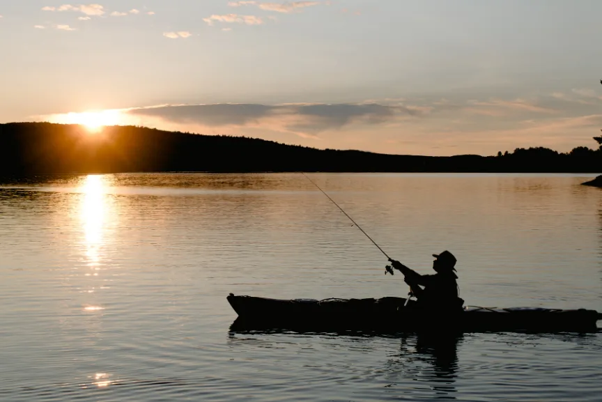 A person in a kayak is fishing on a calm lake during sunset, with the sun low on the horizon and the water reflecting the golden light.