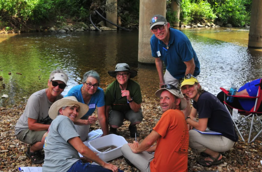 Six adults wearing hats and casual clothing kneel and crouch together by a creek under a bridge, smiling at the camera. They are gathered around a white tray, possibly conducting a nature or science activity.