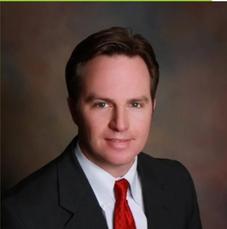 A man with short brown hair wearing a dark suit, white shirt, and red tie poses in front of a brown gradient background.