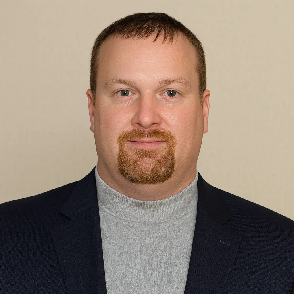 A man with short brown hair and a reddish-brown goatee wearing a gray turtleneck and a dark blazer, facing forward against a plain beige background.