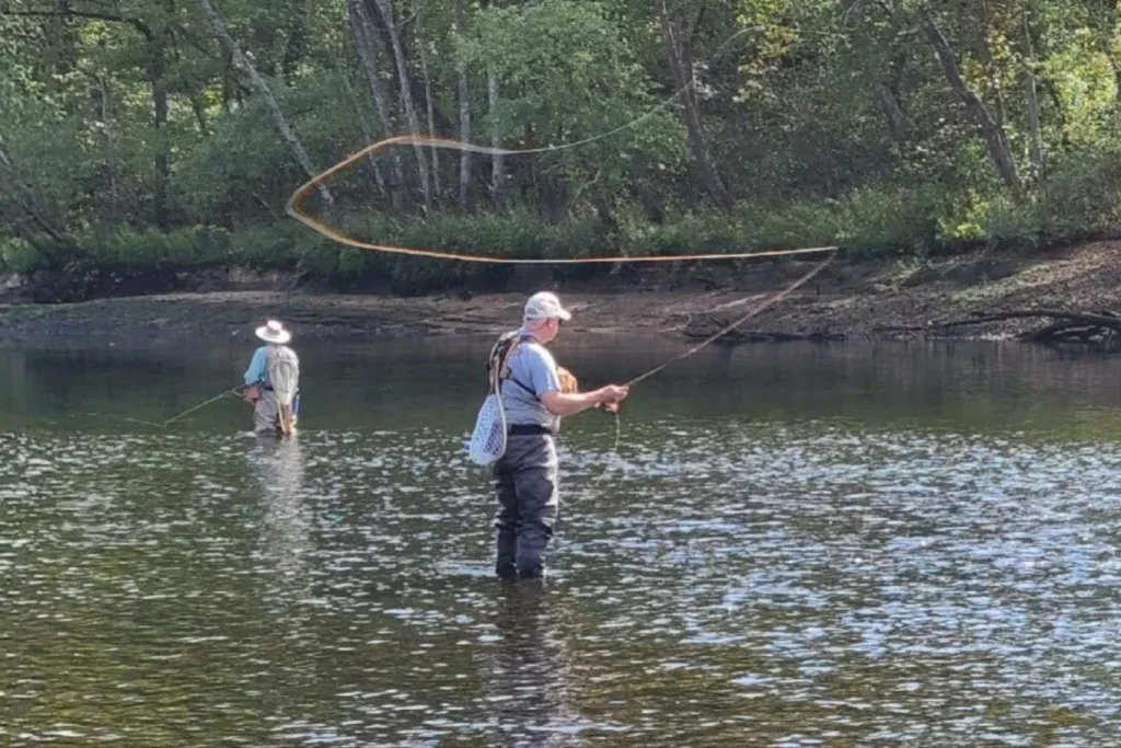 Two people are fly fishing in a shallow river surrounded by trees. One is casting a fishing line, creating a visible loop in the air, while the other stands further back in the water. Both wear hats and waders.