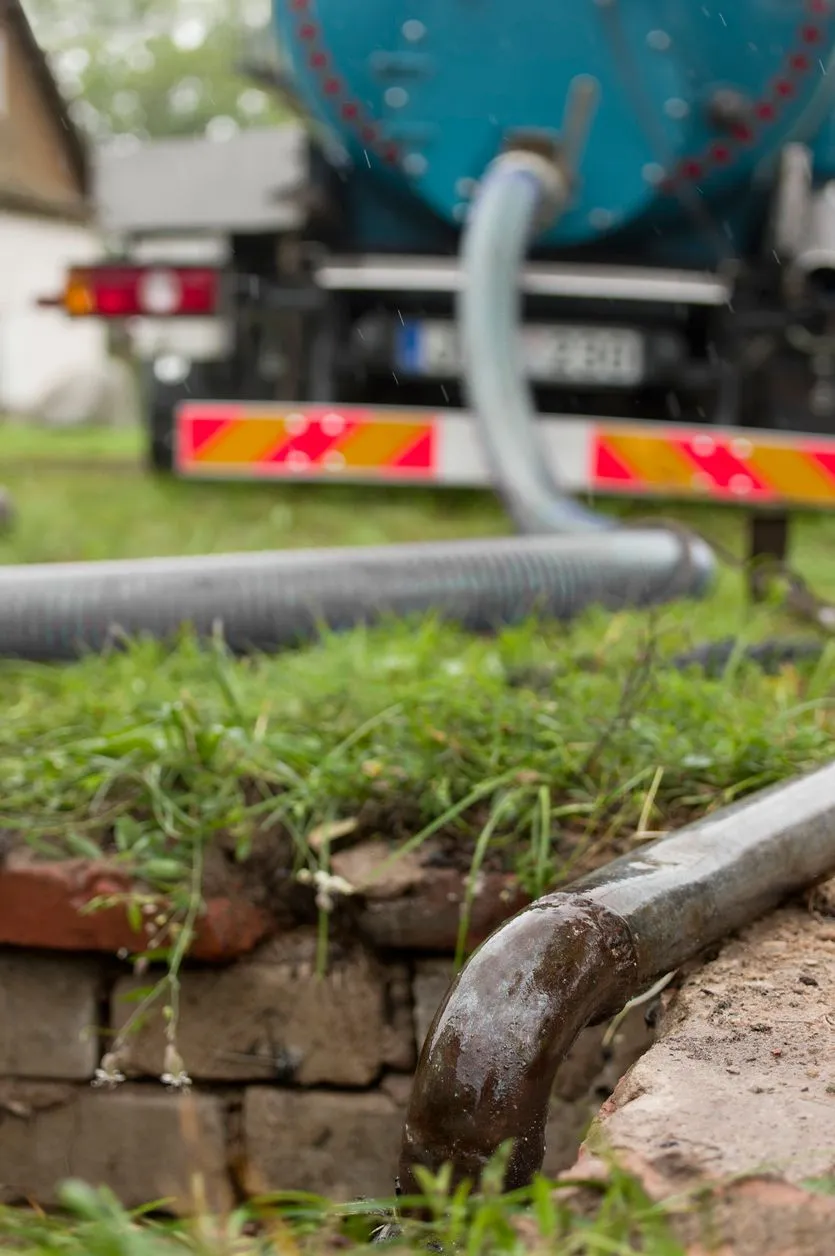 A close-up of a metal pipe and hoses draining liquid from a septic tank, with a blue sanitation truck parked in the background on a grassy lawn.