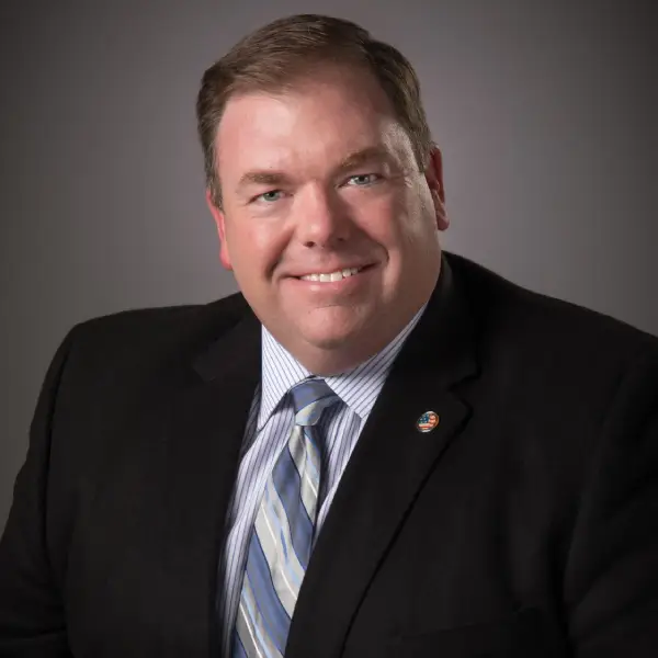 A man in a dark suit and striped tie smiles at the camera against a plain, dark background. He has short brown hair and is wearing a pin on his lapel.
