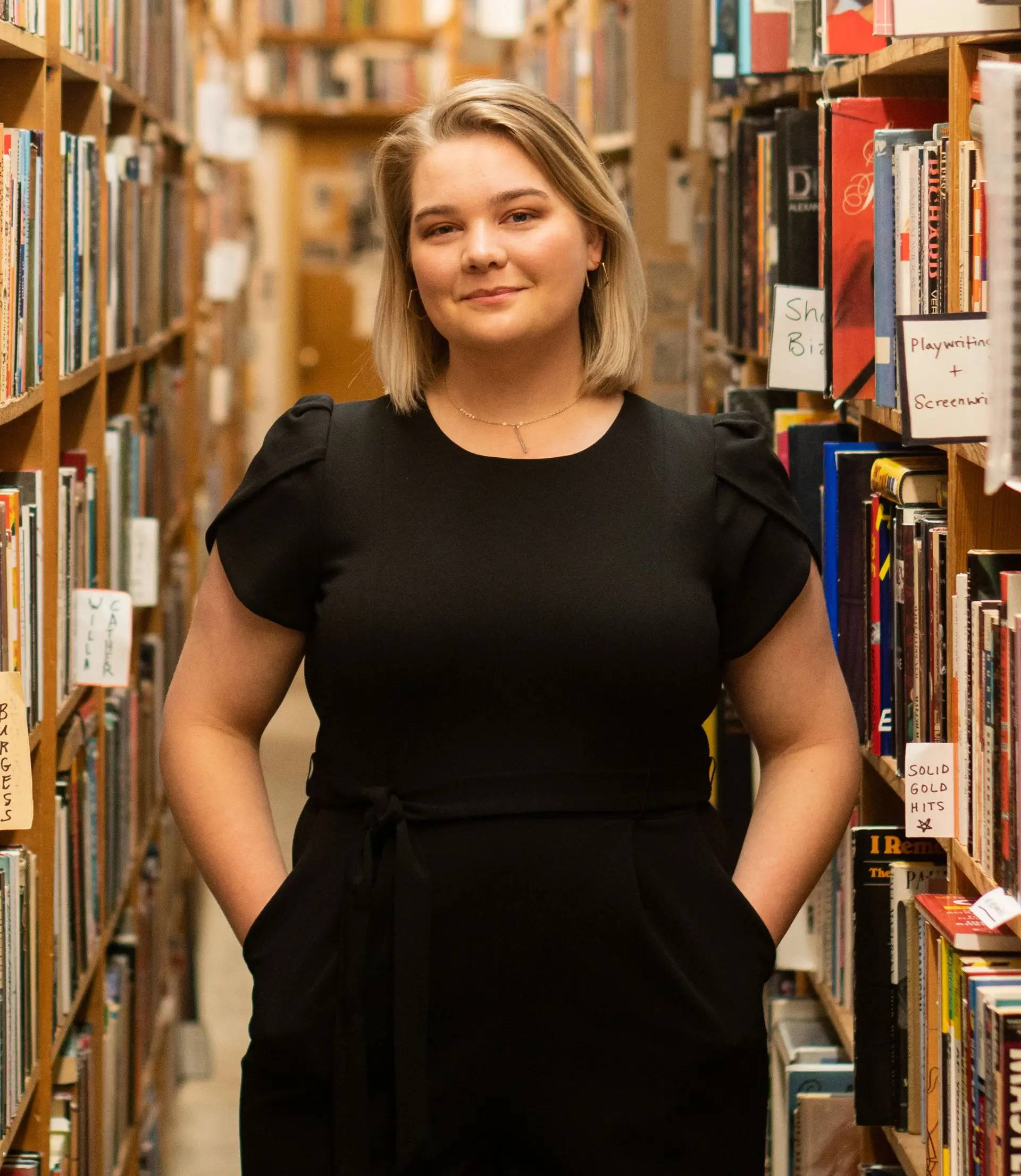A person with blonde hair and a black outfit stands between tall bookshelves in a library, hands in pockets, looking confidently at the camera. The shelves are filled with books and labels.