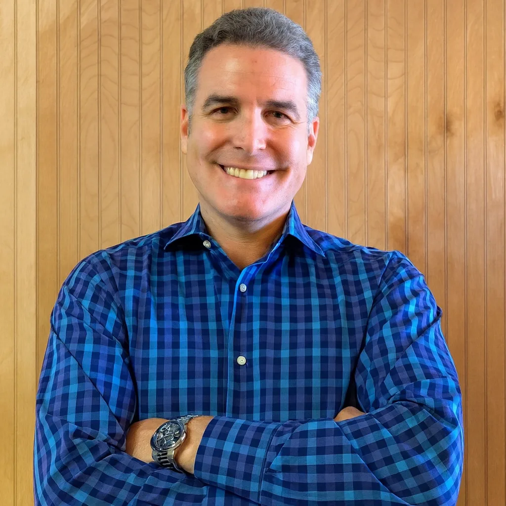 A man with short gray hair, wearing a blue plaid shirt and a wristwatch, stands smiling with arms crossed in front of a light wood-paneled wall.