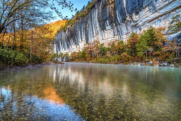 A clear river flows beside tall, layered limestone bluffs, with colorful autumn trees lining the water and rocky cliffs reflecting in the calm surface under a bright blue sky.