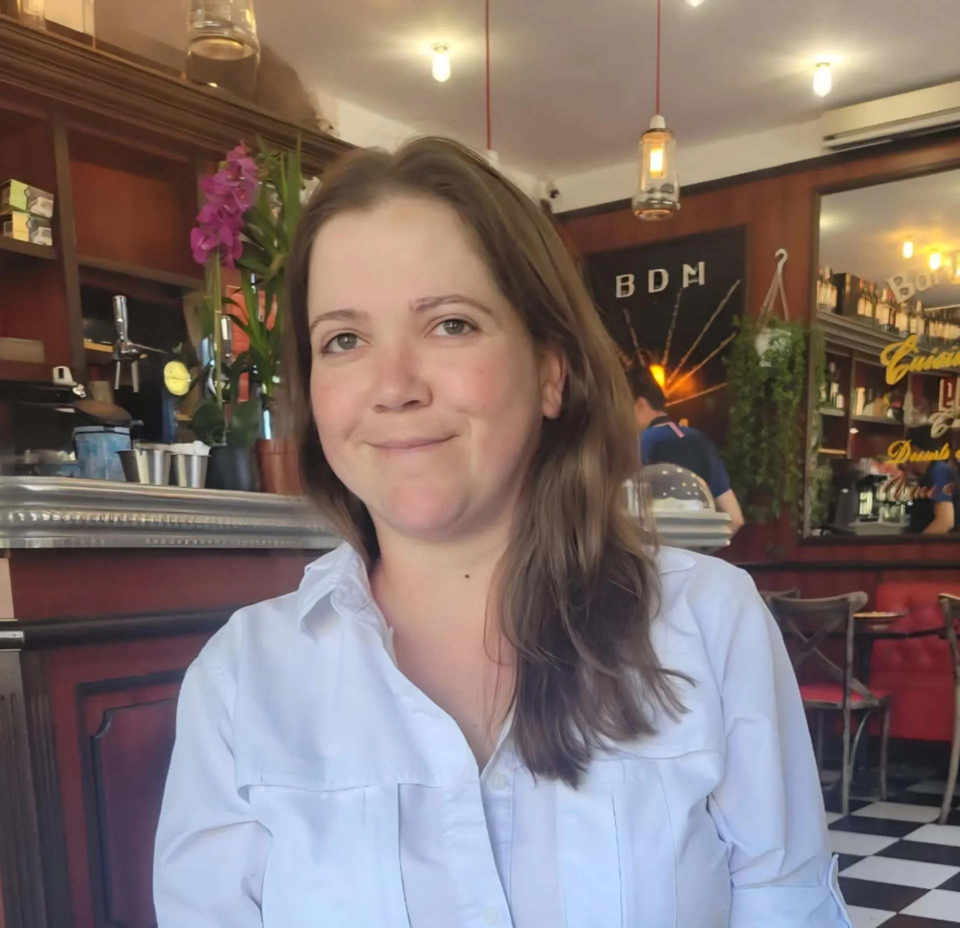 A woman with long brown hair wearing a white shirt smiles while sitting in a cozy café with wooden shelves, plants, and warm lighting in the background.