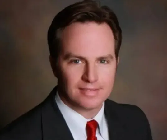 A man in a black suit, blue and white striped tie, and white shirt with a small pin on his lapel.
