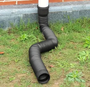A flexible black downspout extension attached to a white gutter pipe directs water away from a brick building, curving across green grass and some small plants.