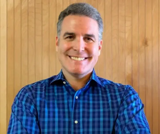 A man wearing a blue plaid shirt smiles at the camera.