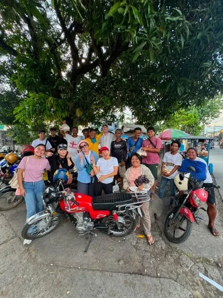 A group of people smile and pose together under a leafy tree on a street, with motorbikes parked nearby. Some hold food items, and the atmosphere appears cheerful and casual.