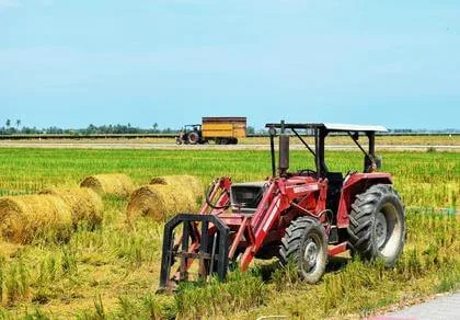 A red tractor sits in a field with hay bales. Another vehicle is seen in the background. The sky is clear and blue, and the field is green and yellow with fresh cut grass.