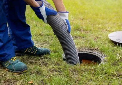 A person in gloves and work boots inserts a large hose into an open outdoor manhole or septic tank, likely performing maintenance or cleaning. Grass and a round cover are visible nearby.