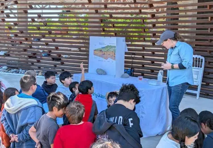 A woman presents what we do in geology to a group of children, some with hands raised, outdoors. A poster with a diagram and rocks sit on a table covered with a white cloth.