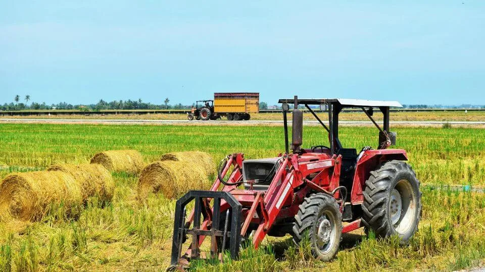 A red tractor parked in a harvested field with hay bales, with another tractor pulling a trailer in the distance.